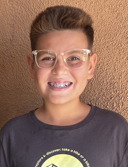 Young person wearing glasses and dark t-shirt smiling at camera against textured tan wall.