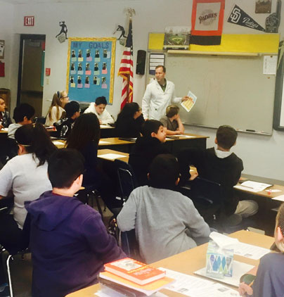 Teacher instructs diverse elementary school students seated at desks in classroom with educational materials displayed.