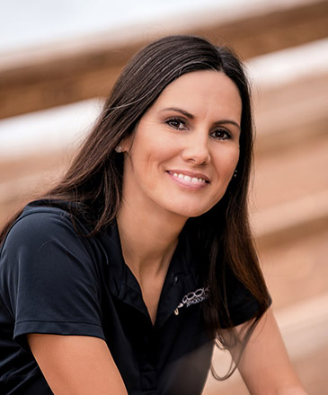 Professional woman smiling at camera wearing black polo shirt, indoor setting with blurred background.
