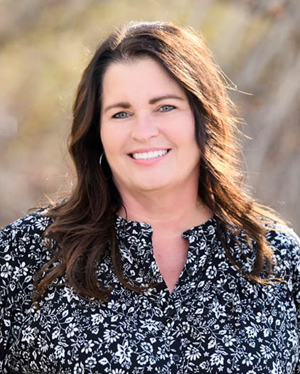Professional headshot of a woman smiling at camera wearing floral patterned blouse, outdoor setting.