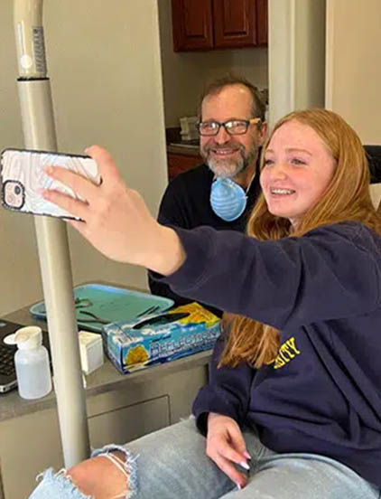 Two people taking a selfie in a kitchen, smiling at phone camera near refrigerator.