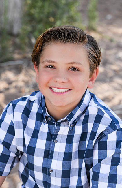 Young boy smiling at camera wearing blue and white checkered shirt outdoors.