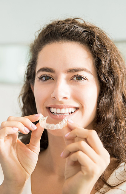 Woman holding clear orthodontic aligners near her smile in a bright clinical setting.