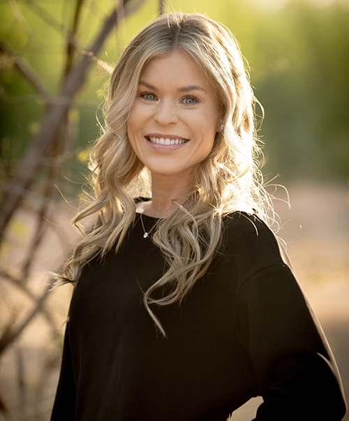 Smiling woman with blonde wavy hair wearing black top outdoors near tree branches.