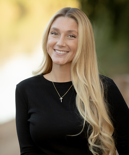 Professional headshot of smiling woman with blonde hair wearing black top and cross necklace.