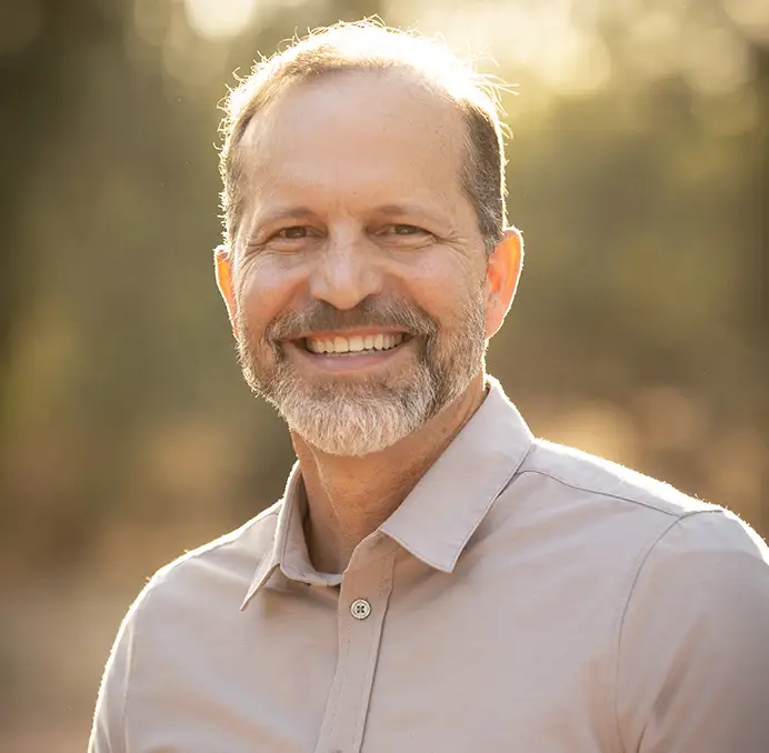 Professional headshot of smiling male orthodontist in light pink shirt, outdoor setting with blurred background.